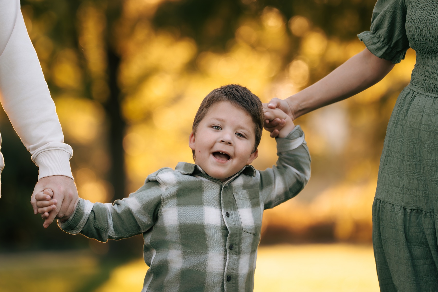 A child laughing while holding an adult’s hand on each side outdoors.