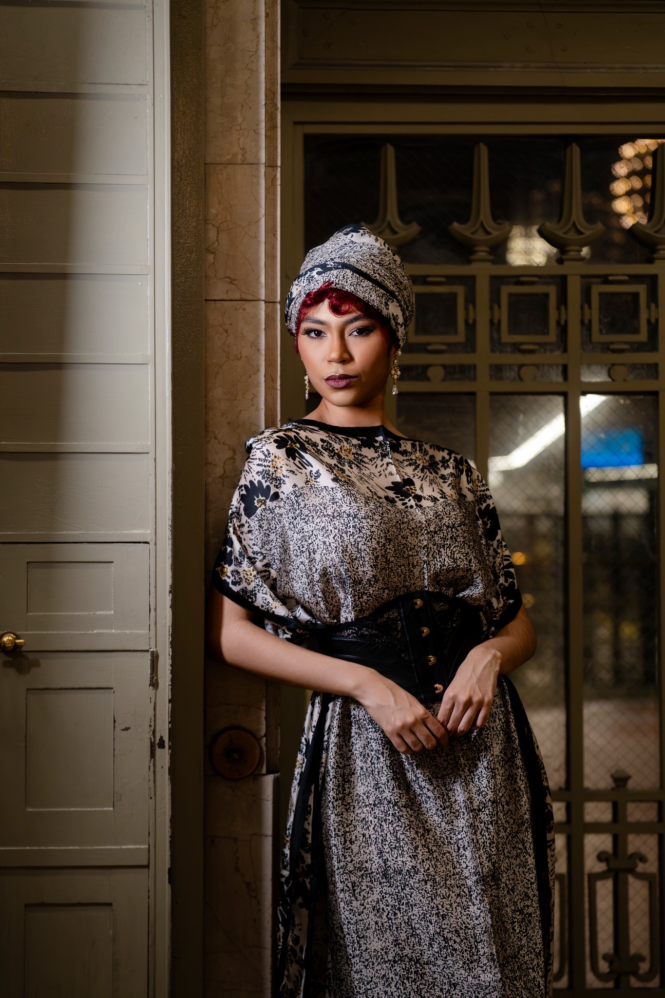 Portrait of a person in a patterned outfit and head wrap framed by an architectural doorway
