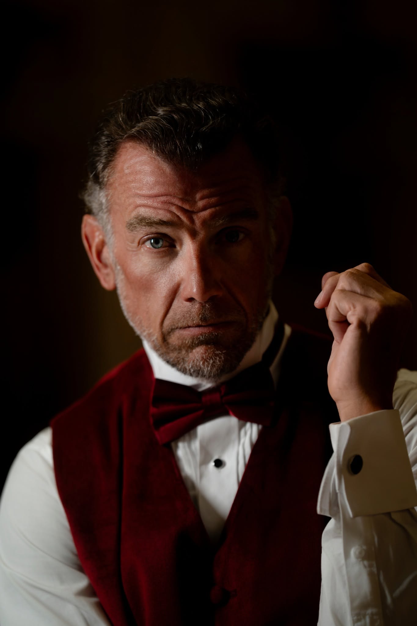 Dramatic close portrait of a man in a burgundy waistcoat and bow tie with light across one side of his face