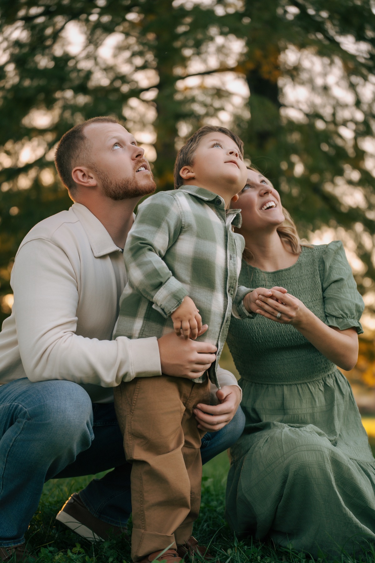 Two adults and a child looking upward together beneath trees.